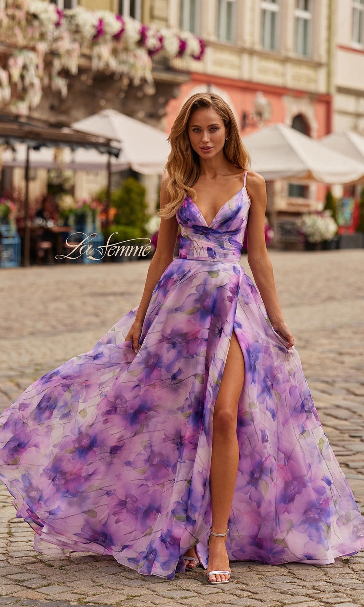 A woman wearing the La Femme 33606 long prom dress in multi stands on a cobblestone street, with outdoor cafes and buildings in the background. The dress features a flowing chiffon fabric, floral print, an a-line skirt, and adjustable spaghetti straps.